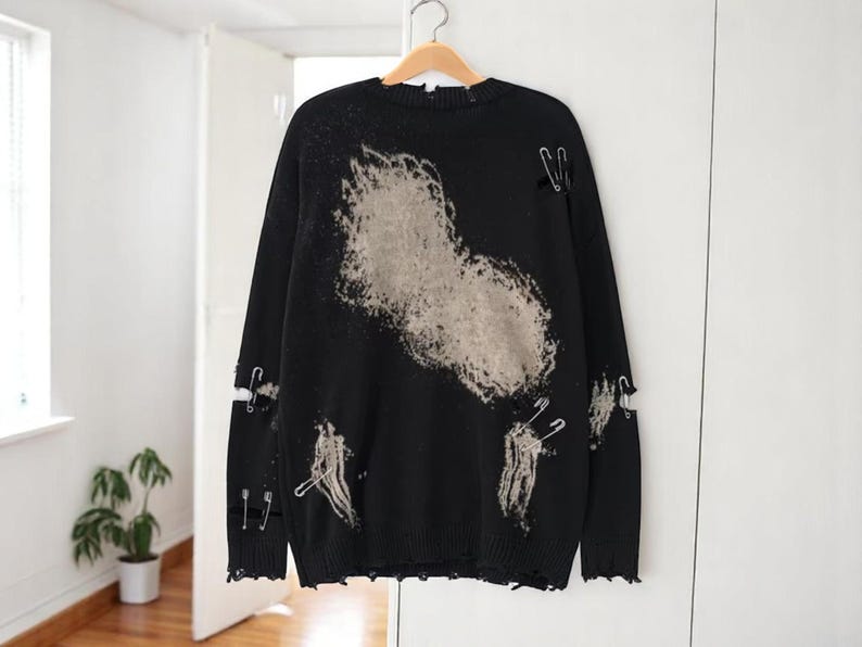 May include: Black distressed sweater with a large, faded beige design on the front. The sweater has ripped details and is adorned with silver safety pins. The sleeves have cut-out details. The sweater is hanging on a wooden hanger.