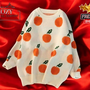 May include: A white knitted sweater with a crew neck and orange embroidered oranges. The sweater has a casual and comfortable look.