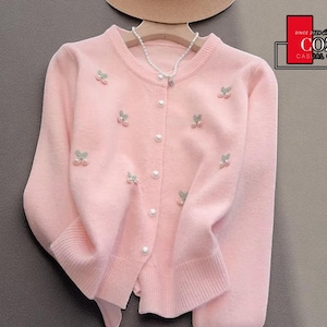 May include: A light pink cardigan sweater with a round neckline and long sleeves. The sweater features pearl buttons and is adorned with embroidered cherry designs. The Cozy Casual Comfort logo is visible in the upper right corner.