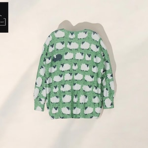 May include: Green sweater with a pattern of white sheep with black faces. The sweater has a crew neck and long sleeves. The Cozy Casual Comfort logo is in the upper left corner.