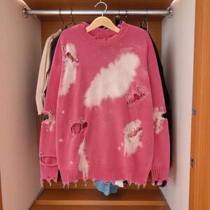May include: A pink distressed sweater with white cloud-like bleach spots hangs in a wooden closet. The sweater has ripped details and is hanging on a wooden hanger. Other clothing items are visible in the background.