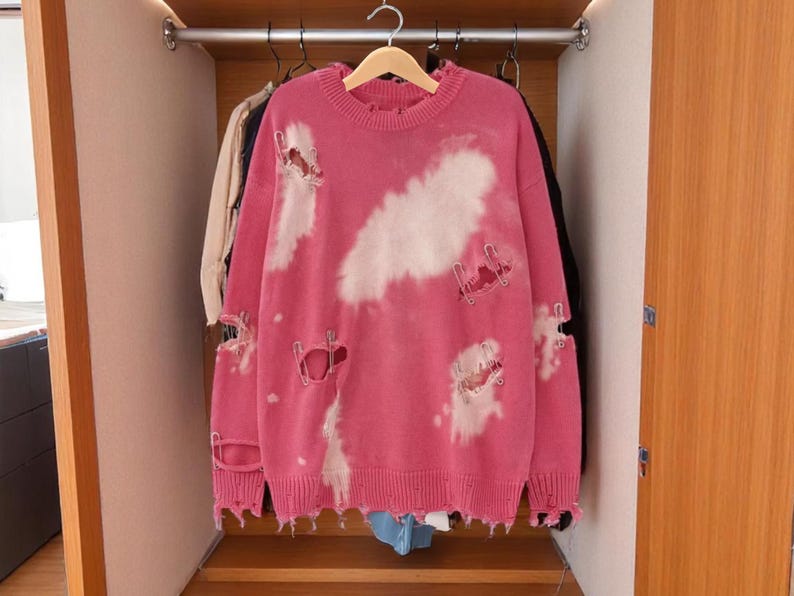 May include: A pink distressed sweater with white cloud-like bleach spots hangs in a wooden closet. The sweater has ripped details and is hanging on a wooden hanger. Other clothing items are visible in the background.