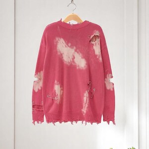 May include: A distressed pink sweater with white cloud-like bleach spots and ripped details. The sweater has safety pins and a frayed hem. It hangs on a wooden hanger against a neutral background.