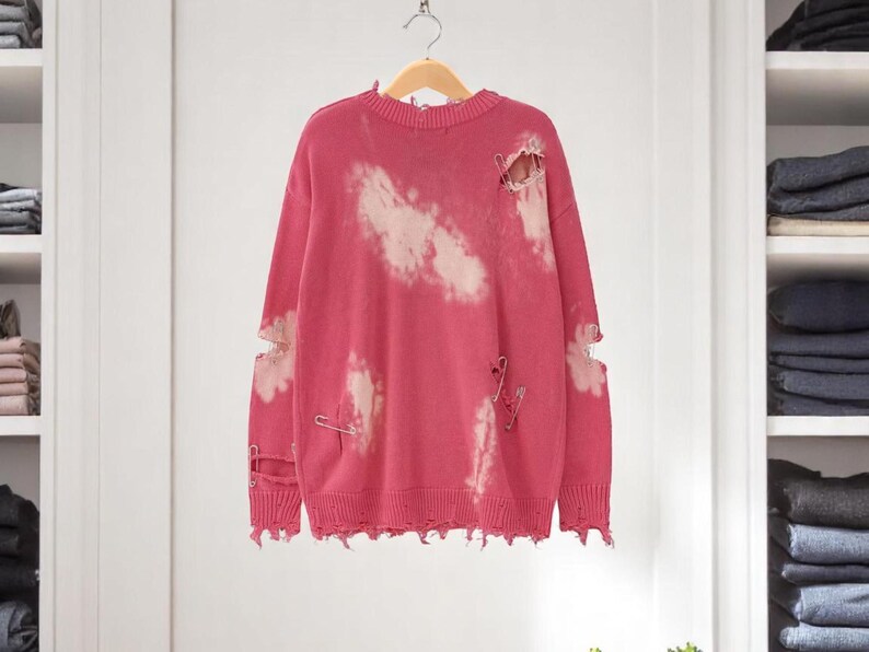 May include: A distressed pink sweater with white cloud-like bleach spots and ripped details. The sweater has safety pins and a frayed hem. It hangs on a wooden hanger against a neutral background.