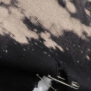 May include: Close-up of distressed black fabric with a bleached, off-white pattern. A tear in the fabric is held together with two silver safety pins. The textile has a worn, grunge aesthetic.