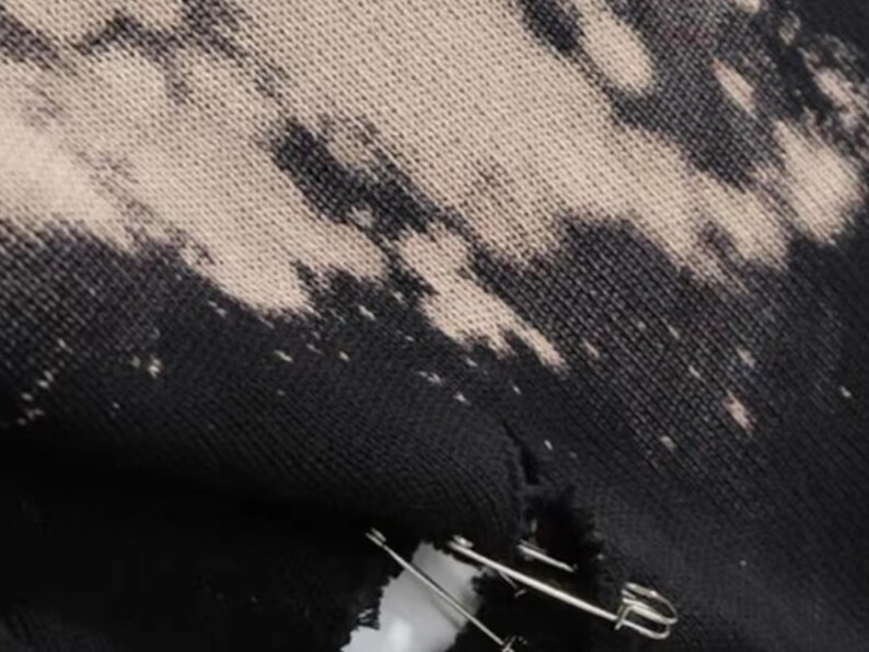 May include: Close-up of distressed black fabric with a bleached, off-white pattern. A tear in the fabric is held together with two silver safety pins. The textile has a worn, grunge aesthetic.