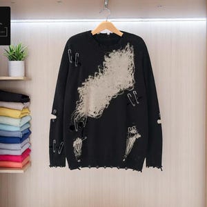 May include: Black distressed sweater with a large off-white design and safety pin accents. The sweater has a crew neck and long sleeves. The Cozy Casual Comfort logo is in the top left corner. The sweater is hanging in a closet.