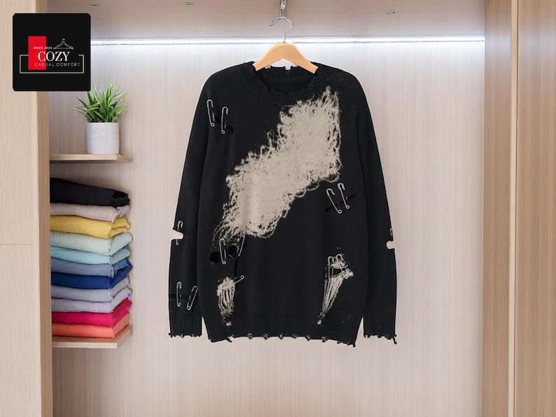 May include: Black distressed sweater with a large off-white design and safety pin accents. The sweater has a crew neck and long sleeves. The Cozy Casual Comfort logo is in the top left corner. The sweater is hanging in a closet.