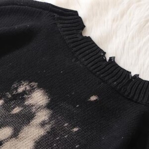 May include: Close-up of a black sweater with a distressed, bleached design. The sweater has a ribbed neckline with a frayed edge. The fabric appears to be a knit material, with a soft, textured appearance. The sweater is likely part of a clothing collection.