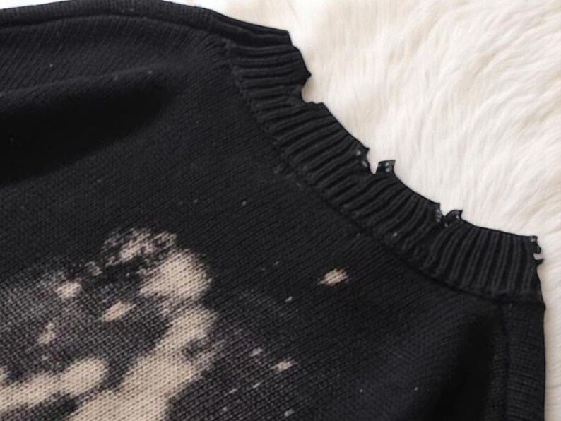 May include: Close-up of a black sweater with a distressed, bleached design. The sweater has a ribbed neckline with a frayed edge. The fabric appears to be a knit material, with a soft, textured appearance. The sweater is likely part of a clothing collection.