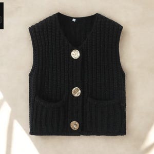 Puede incluir: Un chaleco negro de punto con cuello en V y tres botones dorados decorativos grandes. El chaleco tiene dos bolsillos delanteros y está hecho de un hilo grueso y texturizado. El logotipo "COZY CASUAL COMFORT" está en la esquina superior izquierda.