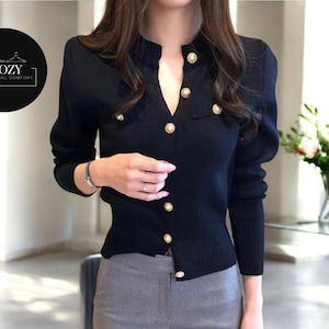 May include: Black cardigan with gold buttons. The V-neck sweater has long sleeves and a ribbed hem. The image includes a logo that says "COZY CASUAL COMFORT". The cardigan is paired with gray pants.