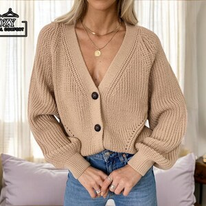 May include: A tan, chunky-knit cardigan with a V-neckline and two dark buttons. The long-sleeved sweater has a relaxed fit and is part of the "COZY CASUAL COMFORT" collection.
