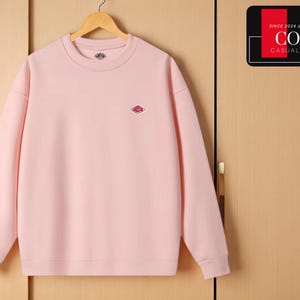 Op de afbeelding: Een lichtroze sweatshirt met een ronde hals en lange mouwen. Het heeft een klein ruitvormig logo op de borst en een bijpassend logo op de halslijn. Het sweatshirt hangt aan een houten hanger. De afbeelding bevat ook een bord met de tekst "COZY CASUAL COMFORT".