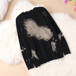 May include: Black distressed sweater with a large, faded gray design on the front. The sweater features ripped details and is adorned with safety pins. The long-sleeved top is displayed on a fluffy white surface.