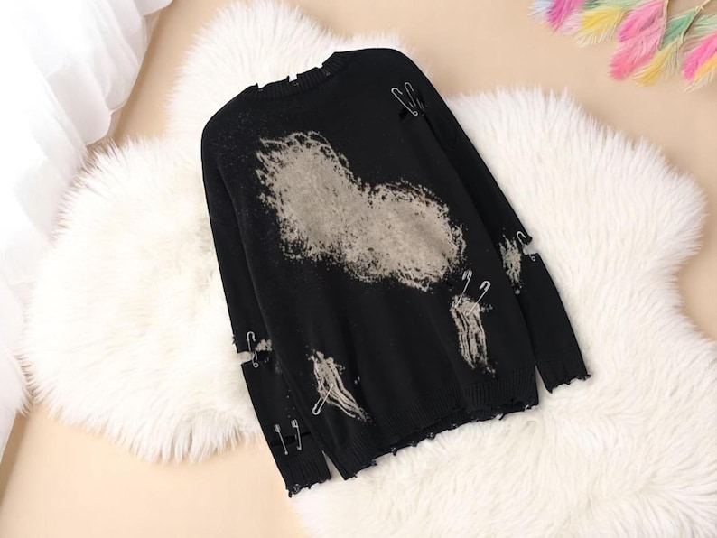 May include: Black distressed sweater with a large, faded gray design on the front. The sweater features ripped details and is adorned with safety pins. The long-sleeved top is displayed on a fluffy white surface.