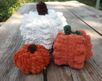 May include: Three crocheted pumpkins, one white and two orange, on a wooden surface. The white pumpkin has a brown stem and the orange pumpkins have green stems.