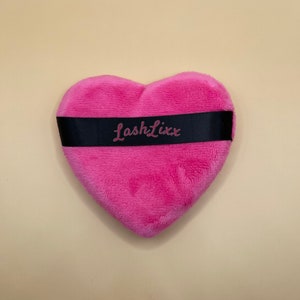 以下が含まれることがあります： ピンクのハート型のメイクアップスポンジ。黒いリボンにピンクの文字で「LashLixx」と書いてあります。