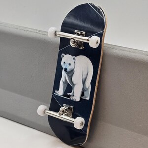 以下が含まれることがあります： 白熊のイラストが描かれた、濃いネイビーブルーのフィンガーボード。シルバーのトラックと白いウィールが付いています。青と白の文字が入った小さなステッカーも写っています。スケートボードのミニチュア版です。
