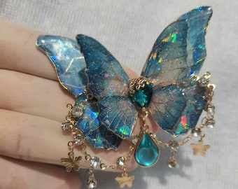 May include: A blue butterfly-shaped brooch with iridescent wings and a teal gemstone center. The brooch features gold-colored accents, dangling chains with clear crystals, and small butterfly charms. The wings have a glittery effect.