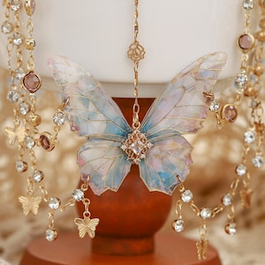 Puede incluir: Un delicado collar de oro con un colgante de mariposa. La mariposa está hecha de material iridiscente y tiene una combinación de colores azul claro y rosa. El collar está adornado con pequeños cristales y tiene una cadena delicada.