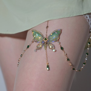 Puede incluir: Una liga de pierna dorada con un diseño de mariposa. La mariposa tiene alas verdes iridiscentes y está adornada con piedras preciosas verdes y transparentes. Una delicada cadena con gemas verdes y transparentes cuelga de la mariposa.