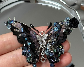 May include: A decorative butterfly brooch with a dark, iridescent wing design. The brooch features intricate details, including small blue and black floral accents, and a silver-toned center with a dangling chain.
