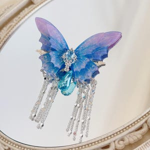 Könnte beinhalten: Blau-violetter Haarclip in Schmetterlingsform mit silbernen Akzenten. Die Flügel haben ein transparentes Aussehen mit filigranen Details. Hängende Silberketten und ein großer blauer Edelstein ergänzen das Design. Auf einer reflektierenden Oberfläche.