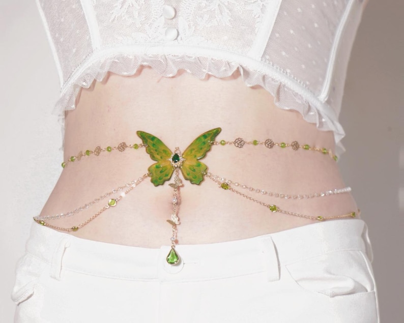 K&ouml;nnte beinhalten: Ein goldener Ketten-K&ouml;rper-Schmuck mit einem gr&uuml;nen Schmetterlings-Anh&auml;nger und gr&uuml;nen Kristall-Akzenten.