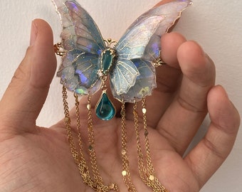 May include: A butterfly-shaped hair clip with iridescent blue and white wings. The clip features a gold-colored frame, a teal-colored gem, and dangling gold chains with small disc accents. The butterfly is held in a hand.