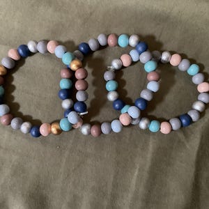May include: Three beaded bracelets in shades of blue, pink, and grey. The bracelets are made of wooden beads and are stacked on top of each other.