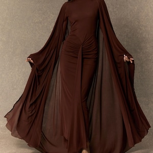 May include: A floor-length dark brown dress featuring a high neckline, long flowing cape-like sleeves, and a gathered, ruched detail at the center waist that creates a draped effect down the front of the skirt, extending to the floor.