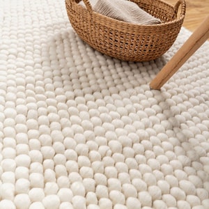 Puede incluir: Una alfombra de color crema construida con numerosas pequeñas bolas de lana afieltrada redondeadas dispuestas en un patrón de cuadrícula denso. Una cesta de mimbre marrón tejida con una tela beige doblada descansa sobre la alfombra.