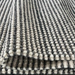 May include: Close-up of a grey and white textured rug. The rug features a repeating pattern of small, raised, rounded bumps, creating a tactile surface. The colour scheme is a neutral combination of grey and white, suitable for various interior styles.