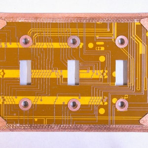 May include: A copper-colored light switch plate with a gold-colored circuit board design. The circuit board has three rectangular cutouts for light switches.