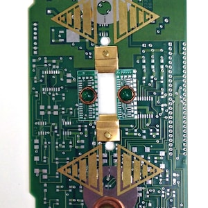 May include: A green circuit board with gold and copper components. The board has a symmetrical design with two triangular shapes at the top and bottom. The text "23386 ASSY" and "SS4100959" is printed on the board.