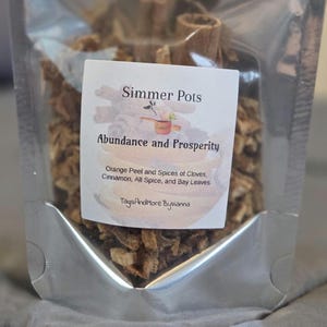 Può includere: Un sacchetto trasparente pieno di spezie ed erbe essiccate. Un'etichetta bianca recita "Simmer Pots Abundance and Prosperity" con ingredienti: scorza d'arancia, chiodi di garofano, cannella, pimento e foglie di alloro. Il marchio "TagsAndMore Byvianna" è visibile.
