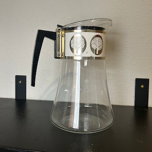 May include: A clear glass coffee pot with a black handle and a silver lid. The pot has a white design with three stylized trees on the side.