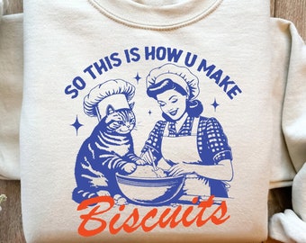 Puede incluir: Una sudadera de color beige claro con un estampado gráfico azul y naranja de un gato y una mujer con gorros de chef haciendo galletas. El texto en la sudadera dice "So this is how u make biscuits".
