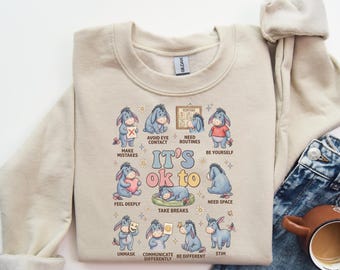 May include: Beige crewneck sweatshirt featuring a graphic of Eeyore from Winnie the Pooh with the text "It's ok to" and various affirmations. The design includes illustrations of Eeyore in different poses and activities.