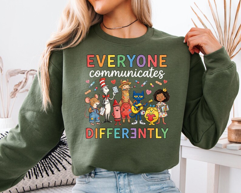 May include: An olive green sweatshirt with the text "EVERYONE COMMUNICATES DIFFERENTLY" in colorful letters. The graphic features cartoon characters including a mouse, cat, llama, and crayon.