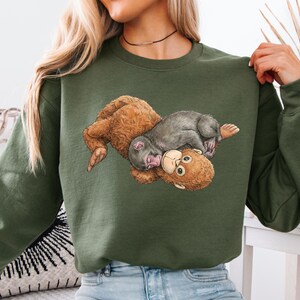 May include: Olive green sweatshirt with a detailed illustration of two monkeys. One monkey is brown, the other is grey, and they are lying on top of each other. The sweatshirt is a solid colour.