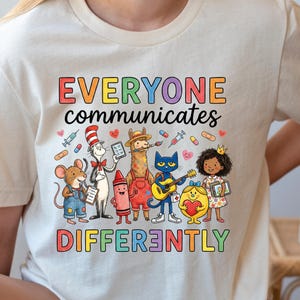 May include: A cream-colored t-shirt featuring the text "EVERYONE communicates DIFFERENTLY" in colorful letters. The graphic includes a mouse, a cat in a hat, a llama, a blue cat with a guitar, a crayon, a yellow character, and a child.