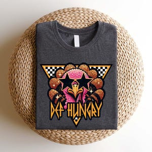 May include: A dark gray t-shirt with a graphic of a pink turkey wearing sunglasses and a star on its head. The turkey is surrounded by gold glitter and the text "DEF HUNGRY" is below it.