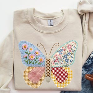 May include: Beige sweatshirt featuring a colorful butterfly applique. The butterfly wings are made of various patterned fabrics, including floral and gingham designs. The butterfly's body is a cross-stitched design. The sweatshirt is folded and placed on a white surface.
