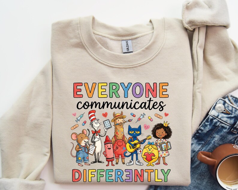 May include: Beige sweatshirt with the text "EVERYONE communicates DIFFERENTLY" in colorful letters. The graphic features cartoon characters including a mouse, the Cat in the Hat, a llama, a cat playing guitar, and a girl with a crown.