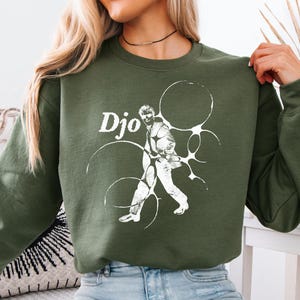 Peut inclure: Un sweat-shirt vert olive avec le mot "Djo" en blanc, et un graphisme blanc d'une personne pratiquant un sport. Le graphisme est entouré de formes circulaires blanches. Le sweat-shirt est en matière souple.