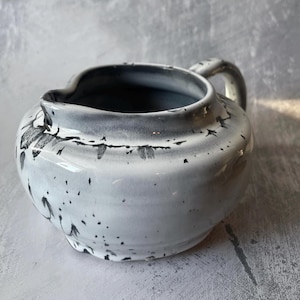 May include: A white ceramic creamer with a black speckled glaze and a handle. The creamer has a rounded shape and a spout for pouring.