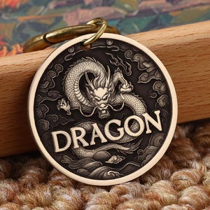 May include: A round, bronze-colored pet tag with a detailed dragon design and the word "DRAGON" in bold letters. The tag is attached to a gold-colored clasp. The background is a wooden surface and a textured rug.