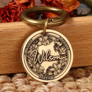 Puede incluir: Una placa redonda dorada para mascotas con el nombre "Wilson" en escritura elegante, rodeada de diseños florales y de hojas. La placa está unida a un cierre color latón. Las palabras "My Love" están inscritas alrededor del borde.
